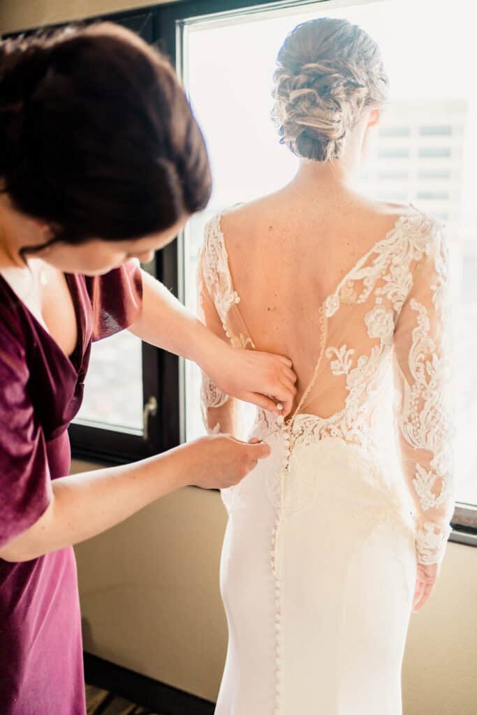 A woman in a wedding dress with intricate lace details is having her dress buttoned by another person. They are near a window with natural light.