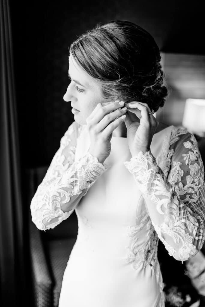 A bride in a white lace gown adjusts her earring, looking out of the frame. The image is in black and white.
