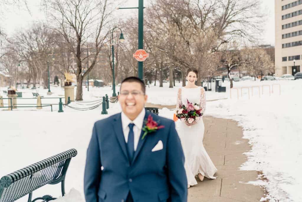 A bride and groom are outdoors in a snowy park. The groom is in focus in the foreground, wearing a navy suit, while the bride in a white dress holding a bouquet is blurred in the background.
