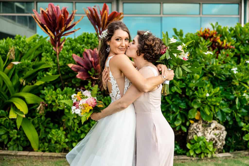 A bride in a white dress and a bridesmaid in a light pink dress share a hug and a kiss on the cheek outdoors, with lush green foliage and red plants in the background.