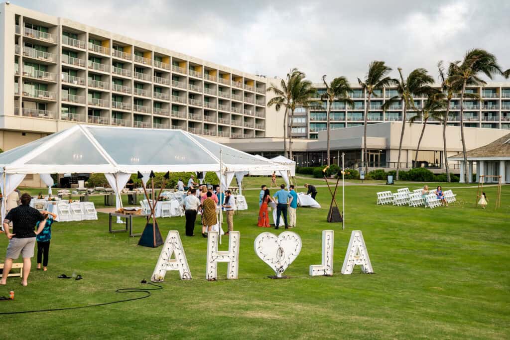 An outdoor gathering with people mingling under clear tents on a lawn in front of a large hotel. Large lit letters spell "Aloha" on the grass. White chairs are arranged for seating.