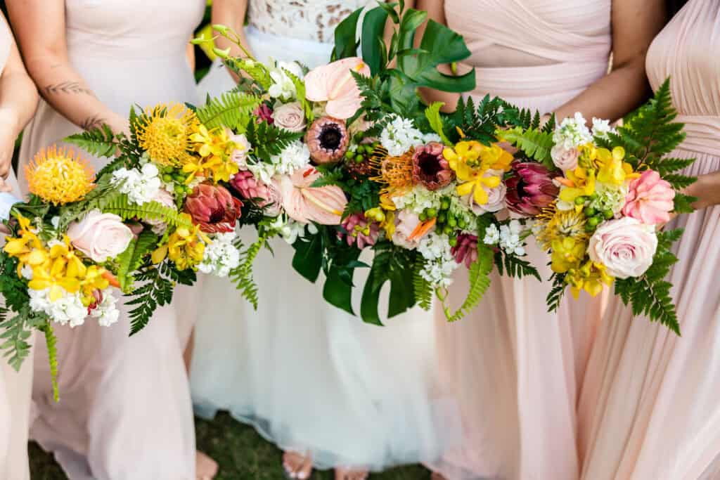 Group of people in pastel dresses holding a vibrant, elongated floral arrangement featuring yellow, pink, white, and green flowers.