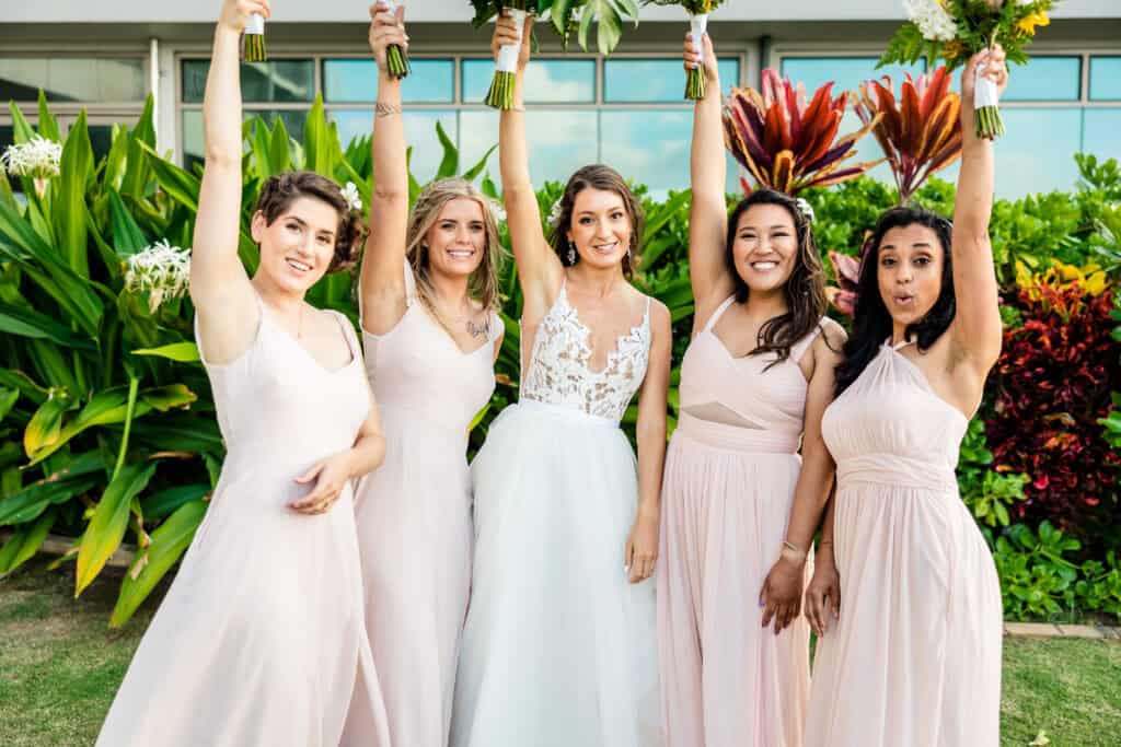 A bride in a white dress poses with four bridesmaids in pink dresses, all holding bouquets up, in front of vibrant greenery and flowers.