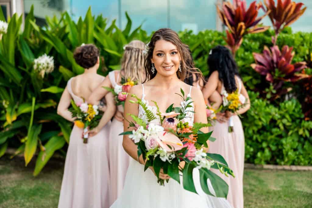 A bride in a white gown holding a bouquet stands in front of bridesmaids in pink dresses, who are facing away, amidst a garden backdrop.
