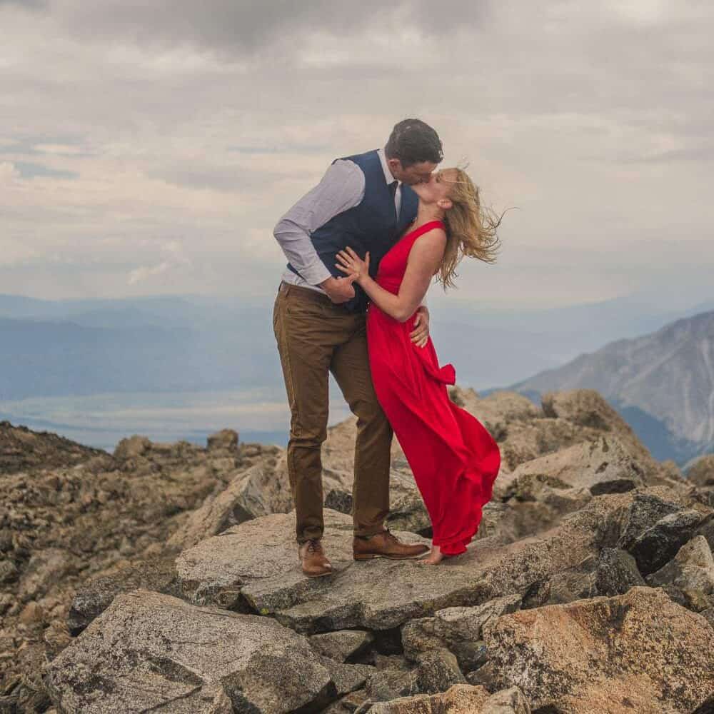 An engaged couple kissing on top of a mountain.