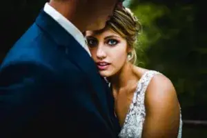 A bride and groom embrace outdoors, captured beautifully by a La Crosse WI Wedding Photographer. The bride looks directly at the camera while resting her head on the groom's shoulder. The groom wears a dark suit; the bride wears a white lace dress.
