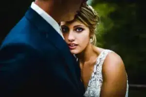 A woman in a white dress rests her head on a man in a dark suit, with greenery in the background.