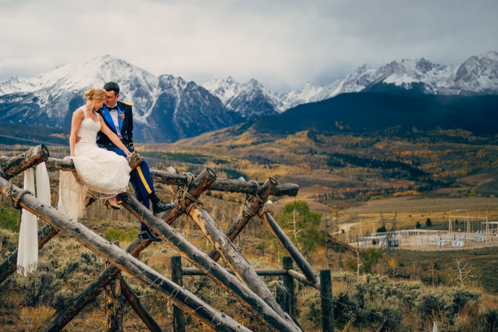 A bride and groom sit on a wooden fence with snow-covered mountains and a vast landscape in the background under a cloudy sky.