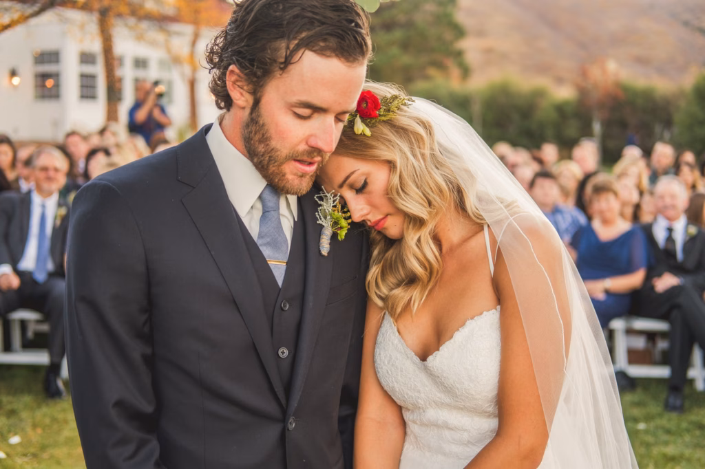 A bride in a white dress and veil rests her head on the groom's shoulder during an outdoor wedding ceremony with guests seated in the background.