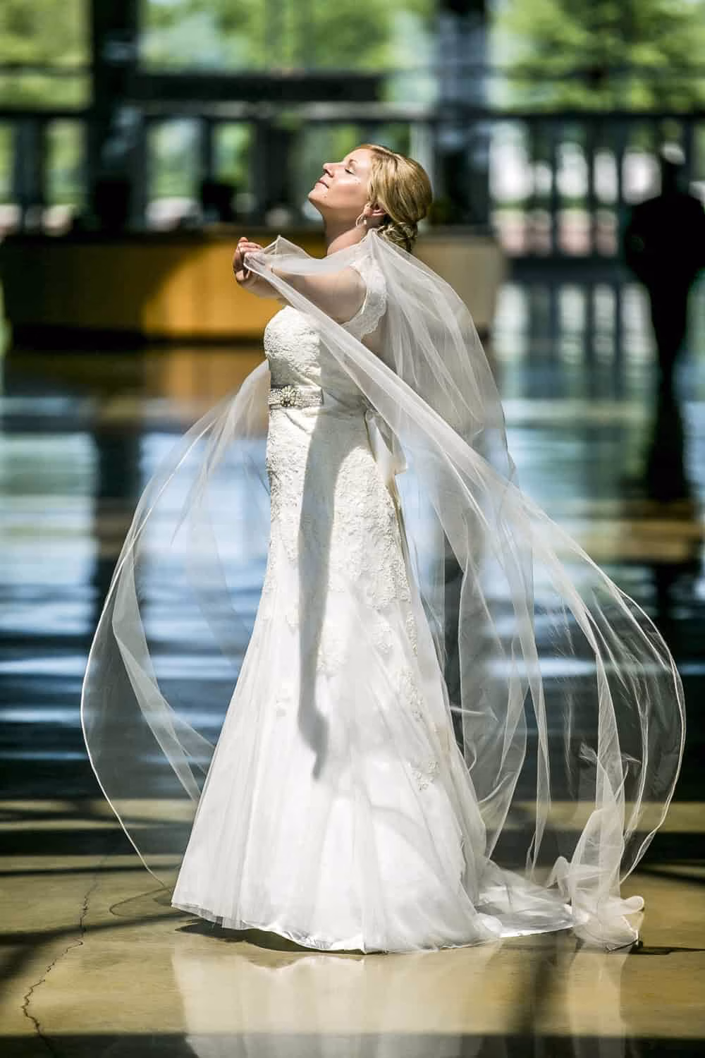 A bride in a white wedding dress stands indoors with her eyes closed, holding her veil as it flows around her in natural light.