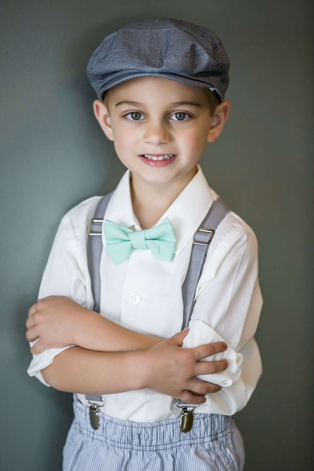 Young boy standing against a gray wall, dressed in a white shirt, mint green bow tie, gray suspenders, and a gray flat cap, with arms crossed and a slight smile.