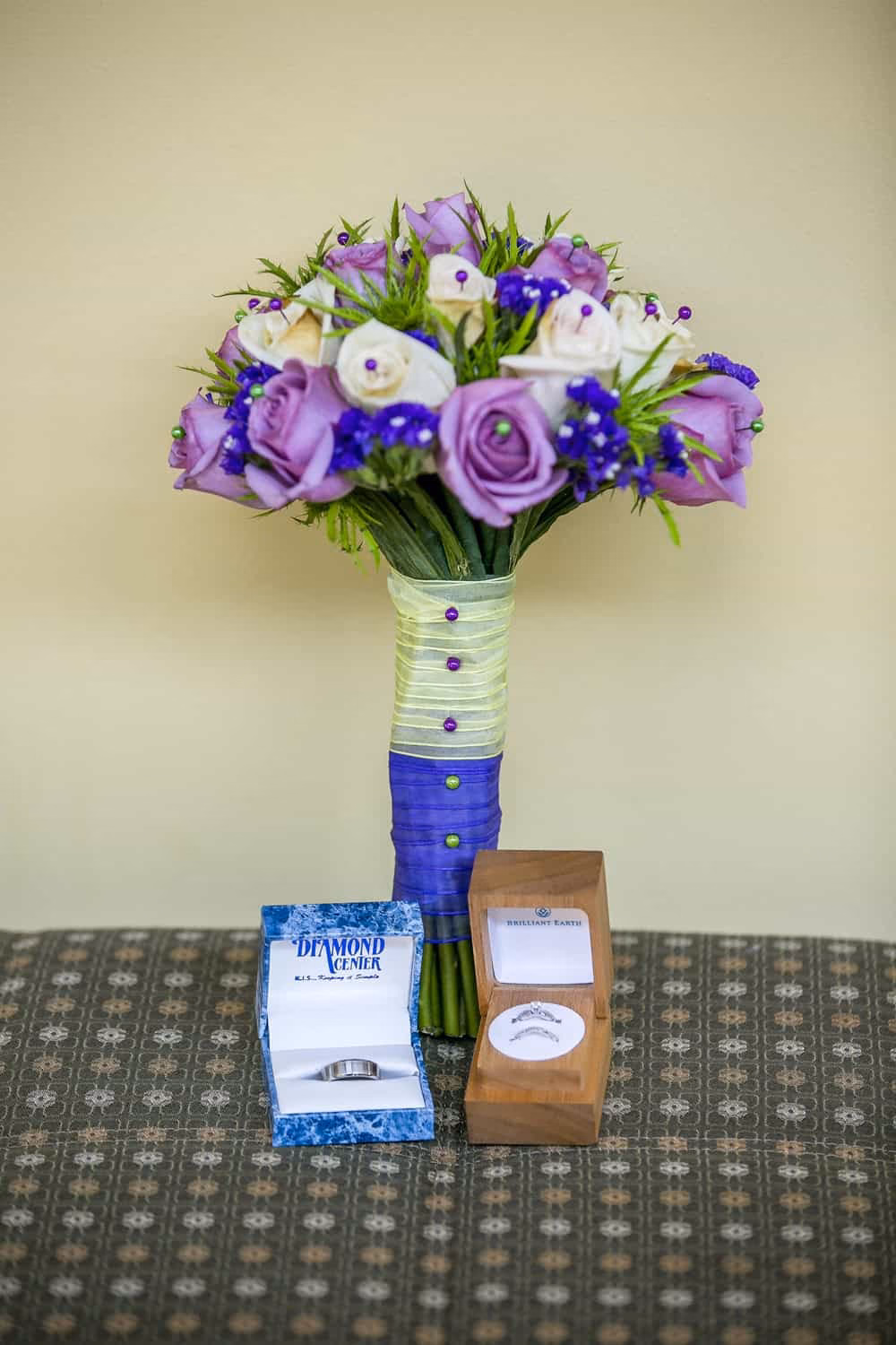 A bouquet of purple and white flowers is displayed on a patterned surface with two ring boxes, one white and one brown, placed in front of it.
