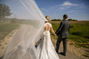 A bride and groom hold hands while walking outdoors on a sunny day, with the bride's veil flowing dramatically in the wind.