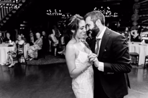 A bride and groom smiling and holding hands as they share their first dance at a wedding reception, beautifully captured by a Vail Wedding Photographer, with guests seated in the background.