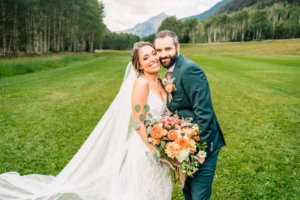 Bride and groom in wedding attire stand close together, smiling and holding a bouquet, with green grass, trees, and mountains in the distance—captured beautifully by a Vail Wedding Photographer.