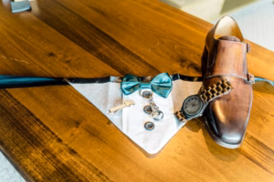 A brown dress shoe, wooden wristwatch, bow tie, vest-shaped napkin, and a small key are arranged on a wooden table—stylishly detailed flatlay by a Vail Wedding Photographer.