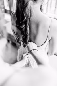 A Vail Wedding Photographer captures a person fastening the back of a woman’s dress as she stands with her back to the camera, her hair styled in loose waves. Black and white photo.