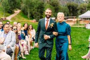 A man in a green suit walks arm-in-arm with a woman in a teal dress down an outdoor aisle, as guests watch and lush greenery sets the scene—captured beautifully by a Vail wedding photographer.