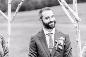 A man in a suit with a boutonniere stands outdoors, smiling slightly next to wooden branches arranged in an arch—captured beautifully by a Vail wedding photographer.