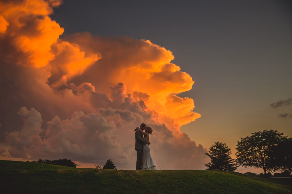 A couple embraces on a grassy hill at sunset, with dramatic orange and purple clouds filling the sky in the background.