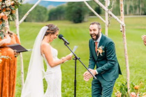 A bride reads vows at an outdoor ceremony, making the groom in a green suit laugh; greenery and floral decorations abound—captured beautifully by a Vail Wedding Photographer.