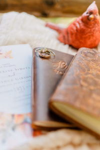 A wedding invitation, two leather-bound vow books with rings on top, and a decorative red cardinal figurine are arranged on a textured surface.