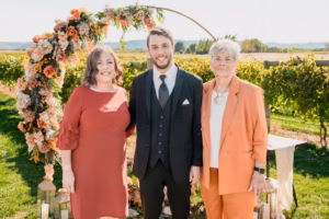 Three people stand outdoors in front of a floral arch at a vineyard, dressed in formal attire, posing for a photo on a sunny day.