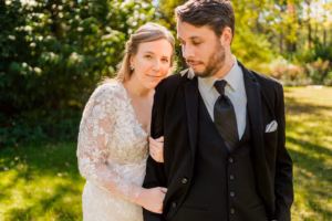 A woman in a white lace dress stands next to a man in a black suit and tie outdoors, with greenery and sunlight in the background.