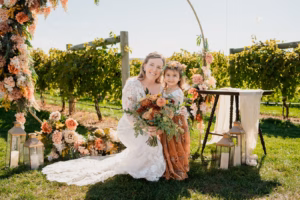 A woman in a white lace wedding dress kneels beside a young girl in a brown dress, both holding a bouquet, surrounded by floral decorations outdoors near a vineyard.