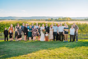 A wedding party of about 25 people poses together outdoors on a green lawn with vineyards and hills in the background under a clear sky.
