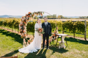 A bride and groom stand together in front of a circular floral arch in an outdoor vineyard setting on a sunny day.