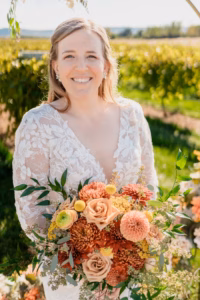 A bride in a white lace dress holds a colorful bouquet of orange, yellow, and pink flowers, standing outdoors in a sunlit garden.