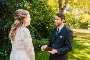 A man in a suit holding a book faces a woman in a white lace dress outdoors, both smiling, with green trees and grass in the background.