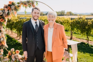 A man in a suit and an older woman in an orange outfit stand smiling together outdoors near a floral arch with vineyards in the background.