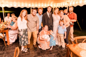 A group of adults and children pose together under a lit tent at an event, with some people smiling at the camera and others holding children.