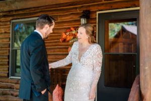 A bride in a lace gown and a groom in a suit smile at each other outside a wooden cabin decorated with a fall floral wreath.