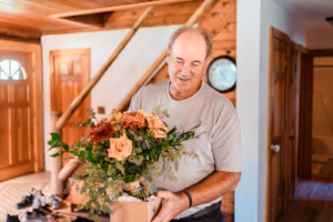 A man in a light gray shirt stands indoors holding a bouquet of flowers with orange and peach blooms.