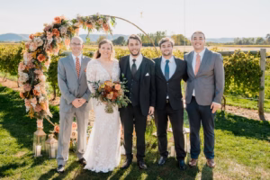 Five people, including a bride in a white gown and four men in suits, stand outdoors in front of a floral arch at a vineyard.