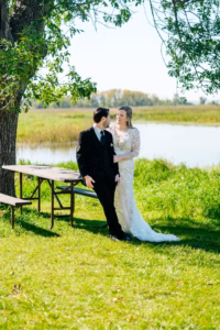 A bride in a white gown stands beside a groom in a black suit by a picnic table near a tree, with a pond and grassy landscape in the background.