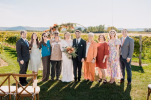 A wedding party poses outside in front of a floral arch, with the bride and groom in the center, surrounded by family and friends.
