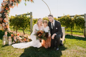 A bride, groom, and young girl kneel and smile outdoors beside a floral arch and lanterns, with green vineyard rows in the background.
