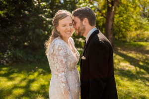 A bride in a lace dress and a groom in a black suit stand close together outdoors, smiling in the sunlight with trees and grass in the background.