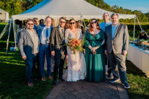 A bride in a white dress and sunglasses stands with six people in formal attire outdoors in front of a white event tent.