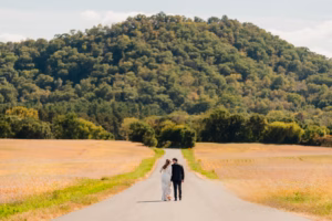 A couple walks down a rural road with fields on both sides and a tree-covered hill in the background.