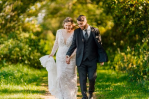 A bride in a white lace dress and a groom in a dark suit walk hand in hand outdoors on a green, sunlit path.