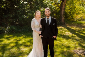 A couple stands outdoors on grass; the woman wears a white lace wedding dress and the man wears a black suit and tie. Trees and sunlight are in the background.