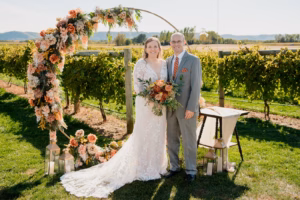 A bride and groom stand outdoors in front of a floral arch, holding a bouquet, with vineyards and hills in the background on a sunny day.