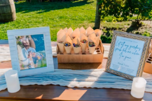 A framed photo, a box of flower petal cones, and a framed sign for a memorial petal toss are displayed on a wooden table outdoors.