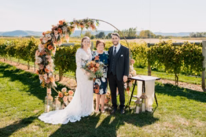 A bride, groom, and an older woman stand in front of a floral arch outdoors at a vineyard wedding ceremony on a sunny day.