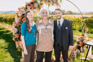 Three people stand in front of a floral arch outdoors, with vineyards and mountains in the background. The group consists of two men and one woman, all smiling at the camera.